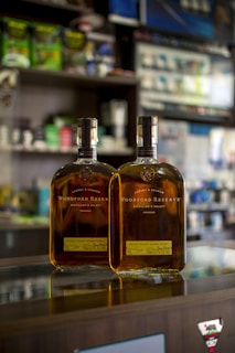 Two bottles of Woodford Reserve bourbon sit on a reflective glass counter in a store. The background features various store shelves with assorted products, slightly out of focus.