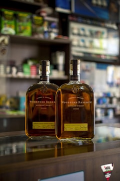 Two bottles of Woodford Reserve bourbon sit on a reflective glass counter in a store. The background features various store shelves with assorted products, slightly out of focus.