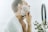 A person is using a razor to shave their face, applying shaving cream and focusing on their neck area. The image includes a reflection in a mirror, which is partially visible along with some green plants in the background.