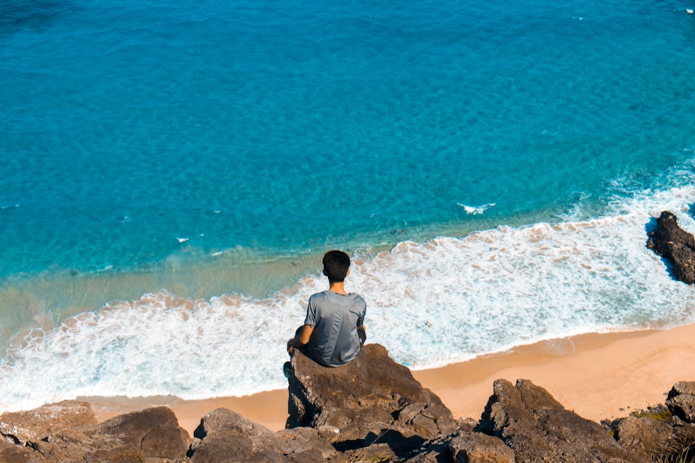 man sitting on top of cliff while looking on seashore
