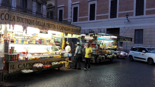 A street market stall in a city selling a variety of products including fruits, drinks, and snacks. The stall is brightly lit with lights, attracting passersby in the evening. Two people appear to be interacting with the vendor while the street is bordered by historic buildings.