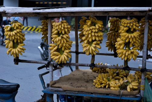 Multiple bunches of ripe bananas are hanging on a wooden cart by the roadside. The scene includes a street setting with a motorcycle and some pedestrians in the background. The bananas are vividly yellow, creating a vibrant contrast against the urban backdrop.