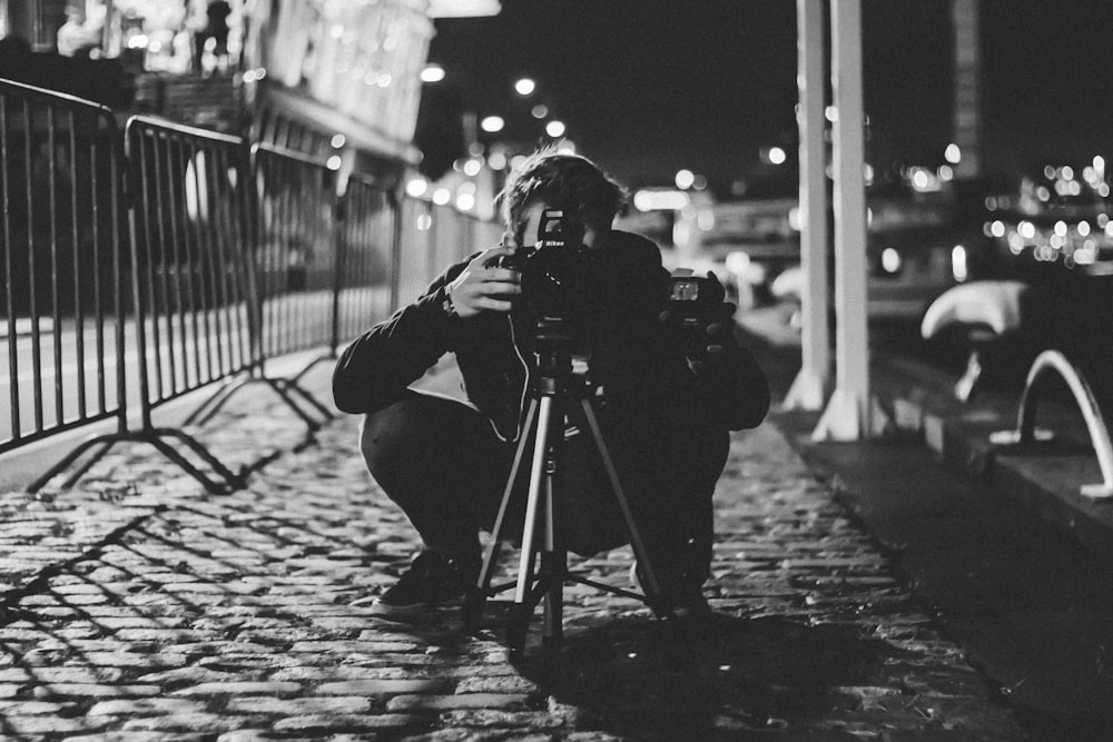 회색 울타리 근처에서 삼각대가 있는 DSLR 카메라를 사용하는 남자의 회색조 사진