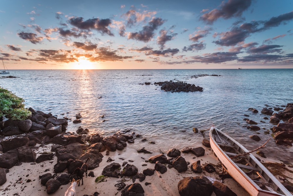 photo of white wooden boat on shoreline near rocks during sunrise