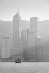 A black-and-white photograph of a cityscape with a traditional-style sailboat on the water in the foreground. The background features towering modern skyscrapers, including buildings with visible logos such as Bank of America Merrill Lynch, AIA, and CCB Asia. The image is slightly hazy, suggesting fog or mist enveloping the city.
