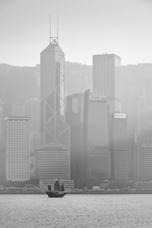 A black-and-white photograph of a cityscape with a traditional-style sailboat on the water in the foreground. The background features towering modern skyscrapers, including buildings with visible logos such as Bank of America Merrill Lynch, AIA, and CCB Asia. The image is slightly hazy, suggesting fog or mist enveloping the city.