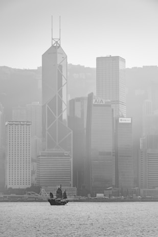 A black-and-white photograph of a cityscape with a traditional-style sailboat on the water in the foreground. The background features towering modern skyscrapers, including buildings with visible logos such as Bank of America Merrill Lynch, AIA, and CCB Asia. The image is slightly hazy, suggesting fog or mist enveloping the city.