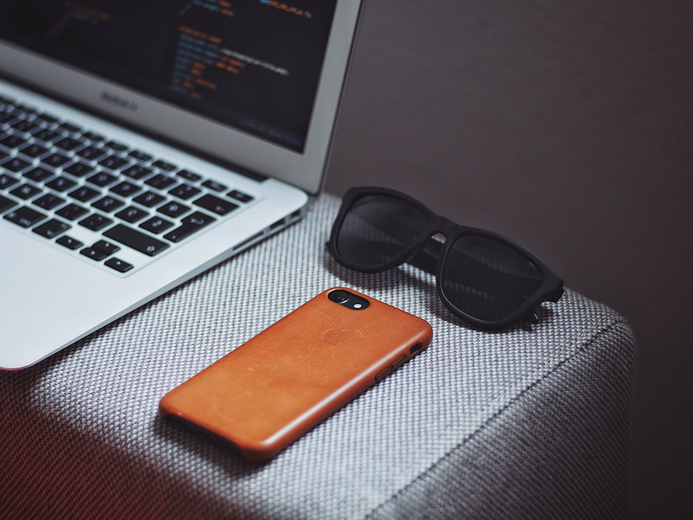 teléfono inteligente naranja cerca de gafas de sol con marco negro al lado de MacBook Air