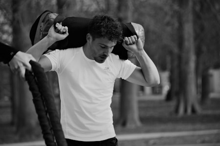 A man in a white athletic shirt is carrying a heavy sandbag on his shoulders while working out in a park. His facial expression shows concentration and effort as he engages in outdoor exercise. The background is blurred, with bare trees indicating it may be autumn or winter.