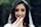 A woman with long dark hair and a veil smiles while holding out a wedding ring in focus in the foreground. The background is blurred with lush greenery, adding a natural and serene setting.