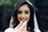 A woman with long dark hair and a veil smiles while holding out a wedding ring in focus in the foreground. The background is blurred with lush greenery, adding a natural and serene setting.