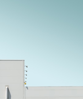 A minimalistic image with pale blue skies and the top part of a white building featuring a few lights protruding from the side and a small yellow cone hanging near the bottom.