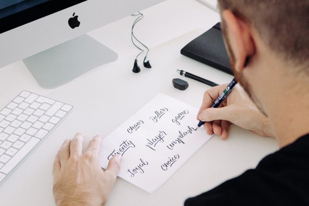 person writing on white printer paper in front of silver iMac