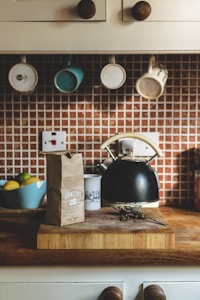 A cozy kitchen scene features a wooden counter with a paper bag labeled 'Atkinsons', a kettle, and a mug. A set of mugs hangs from hooks under a cabinet, with a background of a tiled backsplash. A bowl of fruit, including lemons and limes, sits on the counter to the left.