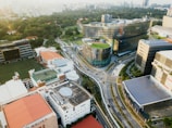 An aerial view of an urban environment featuring modern architecture with a blend of nature. There are prominent buildings with innovative designs, including one with a curved facade and a rooftop garden. Surrounding the structures are well-maintained roads and more rectangular buildings, some with solar panels. Lush greenery is visible in the distance, contributing to a balanced urban landscape.