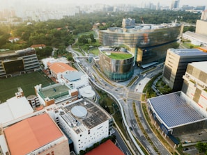 An aerial view of an urban environment featuring modern architecture with a blend of nature. There are prominent buildings with innovative designs, including one with a curved facade and a rooftop garden. Surrounding the structures are well-maintained roads and more rectangular buildings, some with solar panels. Lush greenery is visible in the distance, contributing to a balanced urban landscape.