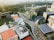 An aerial view of an urban environment featuring modern architecture with a blend of nature. There are prominent buildings with innovative designs, including one with a curved facade and a rooftop garden. Surrounding the structures are well-maintained roads and more rectangular buildings, some with solar panels. Lush greenery is visible in the distance, contributing to a balanced urban landscape.