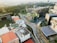 An aerial view of an urban environment featuring modern architecture with a blend of nature. There are prominent buildings with innovative designs, including one with a curved facade and a rooftop garden. Surrounding the structures are well-maintained roads and more rectangular buildings, some with solar panels. Lush greenery is visible in the distance, contributing to a balanced urban landscape.