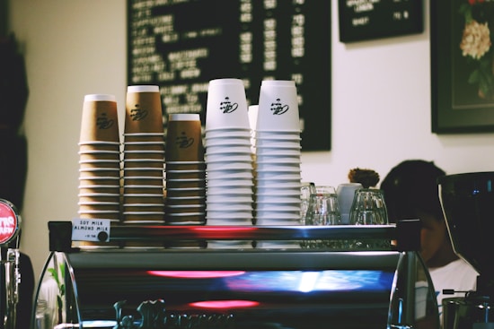 A stack of paper coffee cups, both brown and white, are neatly arranged on top of a coffee machine. Menu boards with lists of coffee options hang on the wall in the background. Various glassware and coffee-making equipment are also visible, contributing to the bustling atmosphere of a caf&eacute; environment.