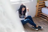 A person with long dark hair, seated on a wooden floor beside a window, holding a book. They are dressed casually in a shirt, vest, tie, blue pants, and sneakers. Light softly illuminates the scene from the window, creating a peaceful atmosphere.