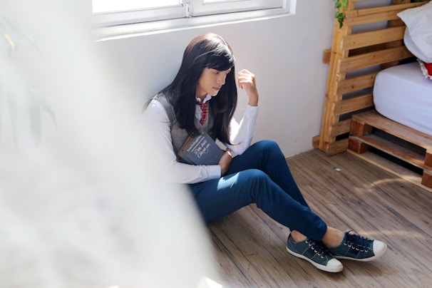 A person with long dark hair, seated on a wooden floor beside a window, holding a book. They are dressed casually in a shirt, vest, tie, blue pants, and sneakers. Light softly illuminates the scene from the window, creating a peaceful atmosphere.