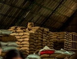 A large pile of burlap sacks is stacked under a corrugated metal roof in a warehouse. The sacks appear to be filled with goods, possibly agricultural products. The lighting is dim, and there is a mix of beige, red, and blue sacks among the mostly brown pile.