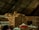 A large pile of burlap sacks is stacked under a corrugated metal roof in a warehouse. The sacks appear to be filled with goods, possibly agricultural products. The lighting is dim, and there is a mix of beige, red, and blue sacks among the mostly brown pile.