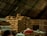 A large pile of burlap sacks is stacked under a corrugated metal roof in a warehouse. The sacks appear to be filled with goods, possibly agricultural products. The lighting is dim, and there is a mix of beige, red, and blue sacks among the mostly brown pile.