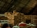 A large pile of burlap sacks is stacked under a corrugated metal roof in a warehouse. The sacks appear to be filled with goods, possibly agricultural products. The lighting is dim, and there is a mix of beige, red, and blue sacks among the mostly brown pile.