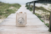 A canvas tote bag with a printed logo is placed on a wooden boardwalk. The boardwalk is surrounded by grass and leads towards a sandy beach area with a view of the sea in the background. The scene has a tranquil, seaside atmosphere.