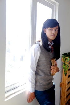 A person with long dark hair stands holding books. They are wearing a white shirt, gray sweater vest, and a red plaid tie, leaning against a window with bright light streaming in from outside. The window is framed by a white wall, and there is a wooden pallet with vines nearby.