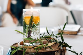 A cylindrical glass candle holder filled with water and containing floating yellow flower petals and green foliage is placed on a wooden slice. Surrounding the candle holder are additional green leaves, creating a natural and fresh look. The setting is likely a table adorned for an event or gathering.