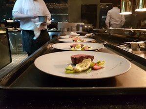 Several plates with artistically arranged food are lined up on a countertop, presumably in a professional kitchen. A chef is partially visible in the background, holding a small pot. Each plate features a piece of seared meat accompanied by vegetables such as asparagus and greens.