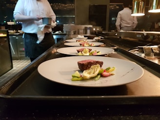 Several plates with artistically arranged food are lined up on a countertop, presumably in a professional kitchen. A chef is partially visible in the background, holding a small pot. Each plate features a piece of seared meat accompanied by vegetables such as asparagus and greens.
