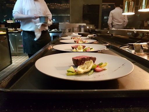 Several plates with artistically arranged food are lined up on a countertop, presumably in a professional kitchen. A chef is partially visible in the background, holding a small pot. Each plate features a piece of seared meat accompanied by vegetables such as asparagus and greens.
