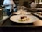 Several plates with artistically arranged food are lined up on a countertop, presumably in a professional kitchen. A chef is partially visible in the background, holding a small pot. Each plate features a piece of seared meat accompanied by vegetables such as asparagus and greens.