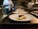 Several plates with artistically arranged food are lined up on a countertop, presumably in a professional kitchen. A chef is partially visible in the background, holding a small pot. Each plate features a piece of seared meat accompanied by vegetables such as asparagus and greens.