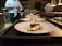 Several plates with artistically arranged food are lined up on a countertop, presumably in a professional kitchen. A chef is partially visible in the background, holding a small pot. Each plate features a piece of seared meat accompanied by vegetables such as asparagus and greens.