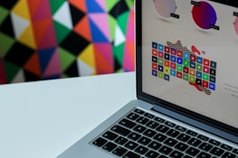 A laptop displaying a colorful grid of app icons is set against a vivid, geometric patterned background. The laptop screen shows various colored circles and app logos, suggesting a focus on digital or graphic design.