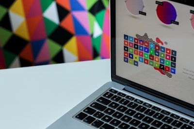 A laptop displaying a colorful grid of app icons is set against a vivid, geometric patterned background. The laptop screen shows various colored circles and app logos, suggesting a focus on digital or graphic design.