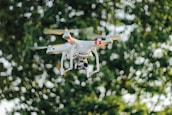A white quadcopter drone equipped with a camera hovers in front of a blurred background of lush green foliage. The drone is captured in mid-flight, with its propellers creating a slight motion blur.