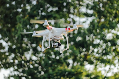A white quadcopter drone equipped with a camera hovers in front of a blurred background of lush green foliage. The drone is captured in mid-flight, with its propellers creating a slight motion blur.