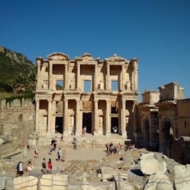 An ancient Roman library ruin featuring a grand facade with multiple columns and arches. Several tourists are exploring the site, surrounded by scattered stones and fragments. The background includes a mountain and clear blue sky.
