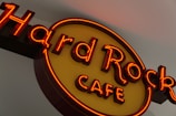 A neon sign with red and yellow colors displaying the words 'Hard Rock Cafe'. The letters are bold and brightly illuminated, creating a vibrant glow against a neutral background.