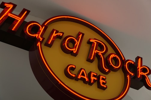 A neon sign with red and yellow colors displaying the words 'Hard Rock Cafe'. The letters are bold and brightly illuminated, creating a vibrant glow against a neutral background.
