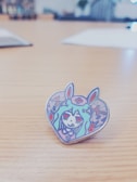 A cartoonish heart-shaped enamel pin featuring an illustration of a character with long hair, wearing a headband with a cross symbol, and bunny ears. The character has red eyes and is surrounded by colorful abstract patterns.