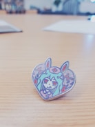 A cartoonish heart-shaped enamel pin featuring an illustration of a character with long hair, wearing a headband with a cross symbol, and bunny ears. The character has red eyes and is surrounded by colorful abstract patterns.