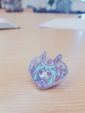 A cartoonish heart-shaped enamel pin featuring an illustration of a character with long hair, wearing a headband with a cross symbol, and bunny ears. The character has red eyes and is surrounded by colorful abstract patterns.