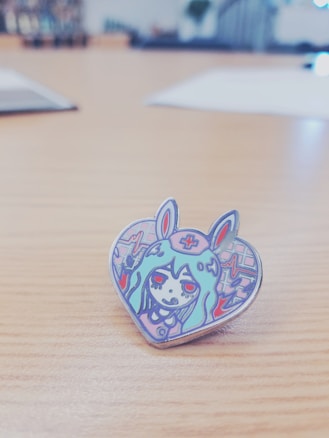 A cartoonish heart-shaped enamel pin featuring an illustration of a character with long hair, wearing a headband with a cross symbol, and bunny ears. The character has red eyes and is surrounded by colorful abstract patterns.