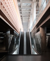 An indoor escalator ascends between two levels, with modern architectural features including glass railings and concrete pillars. The ceiling above is made of beams, providing a sense of depth and openness.